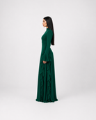 A woman in profile wearing a long-sleeved, high-neck, floor-length emerald green dress with a ruffled skirt, standing against a light gray studio background.