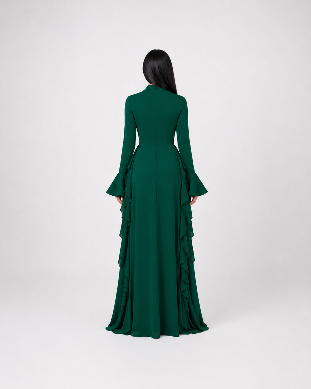 A woman viewed from the back wearing a long-sleeved emerald green gown with a structured waist and layered ruffle detailing along the skirt, set against a plain light gray studio background.