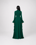 A woman viewed from the back wearing a long-sleeved emerald green gown with a structured waist and layered ruffle detailing along the skirt, set against a plain light gray studio background.