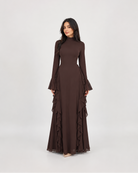 A woman modeling a long-sleeved, high-neck maxi dress in a solid dark brown color, featuring flared bell sleeves and a tiered ruffle skirt, set against a plain light gray studio background.