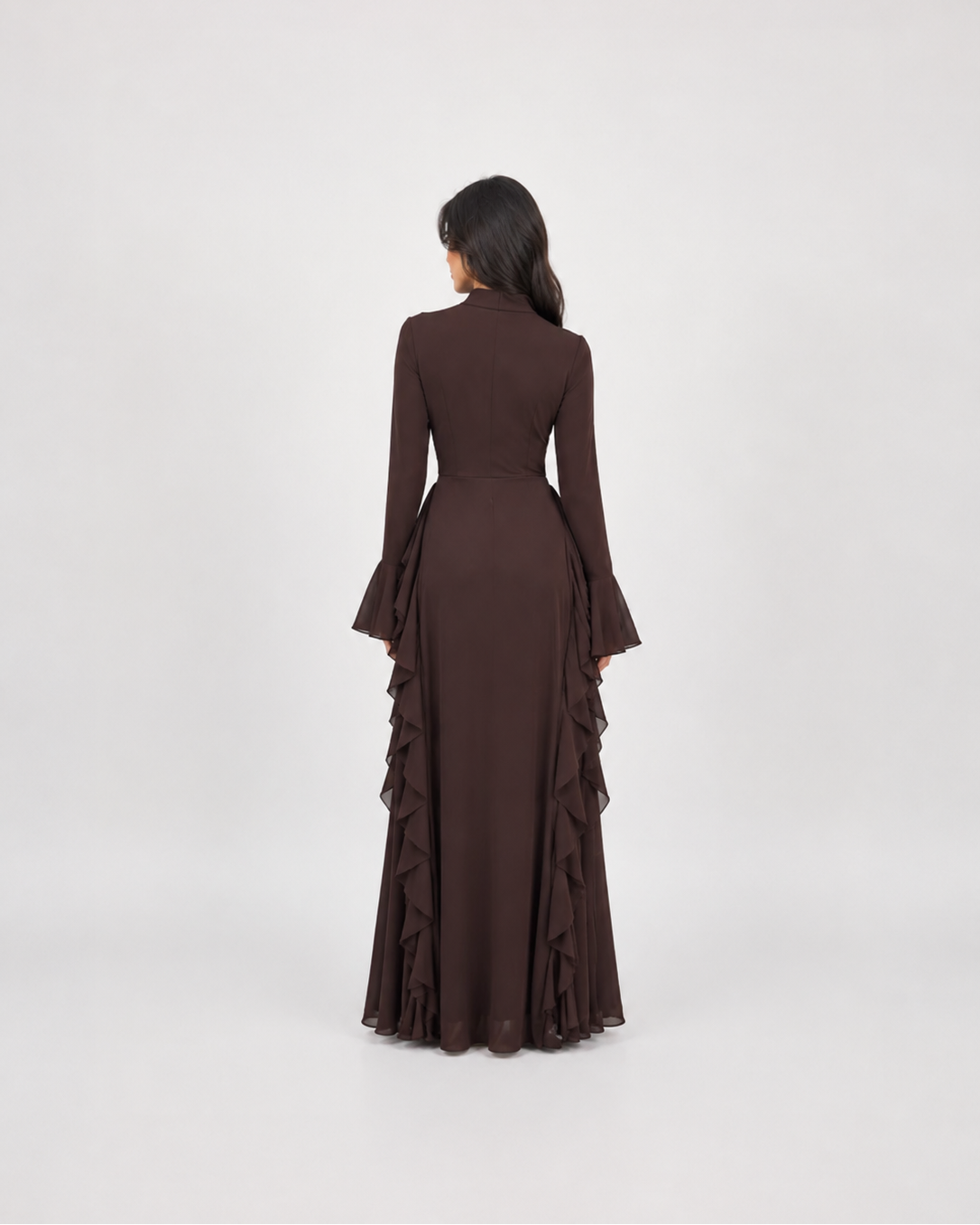 A woman viewed from behind wearing a long-sleeved dark brown maxi dress with flared cuffs and layered ruffle detailing along the skirt, set against a plain light gray studio background.