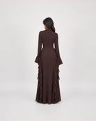 A woman viewed from behind wearing a long-sleeved dark brown maxi dress with flared cuffs and layered ruffle detailing along the skirt, set against a plain light gray studio background.