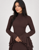 A woman wearing a dark brown long-sleeve mock neck dress with flared cuffs and a tiered ruffle hem, photographed against a light gray studio background.