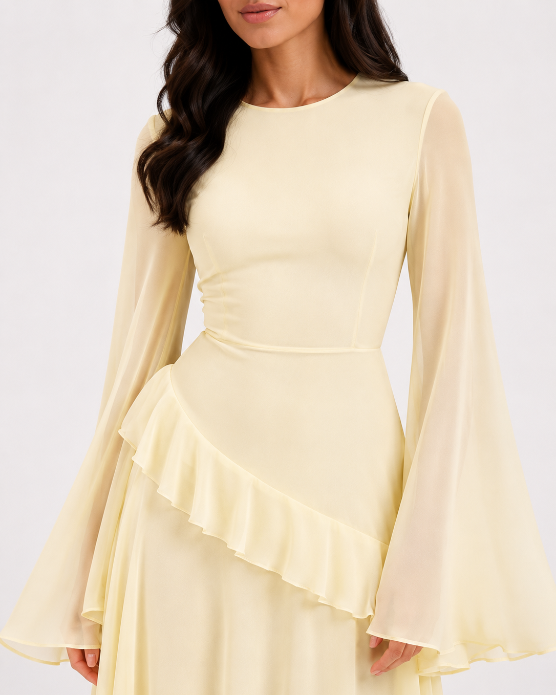 A woman wearing a pale yellow long-sleeve dress with a fitted bodice, ruffled waist detail, and flowing sheer bell sleeves against a plain light background.