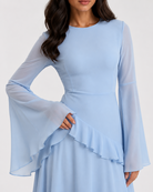 A woman wearing a light blue long-sleeve dress with sheer flared bell sleeves, a fitted waist, and a ruffled overlay detail on the skirt, shown against a plain light background.