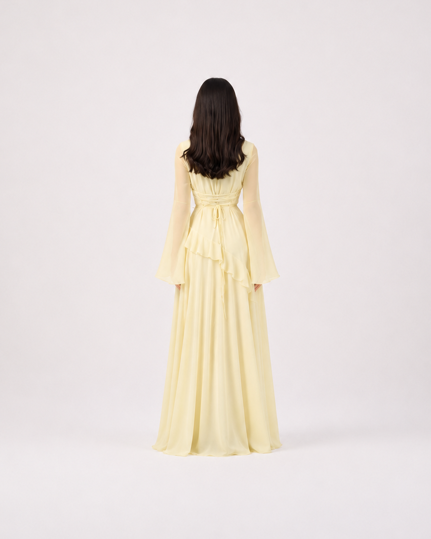 A woman viewed from behind wearing a pale yellow long-sleeve maxi dress with a ruffled waist, standing against a plain light gray studio background.