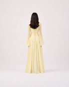 A woman viewed from behind wearing a pale yellow long-sleeve maxi dress with a ruffled waist, standing against a plain light gray studio background.