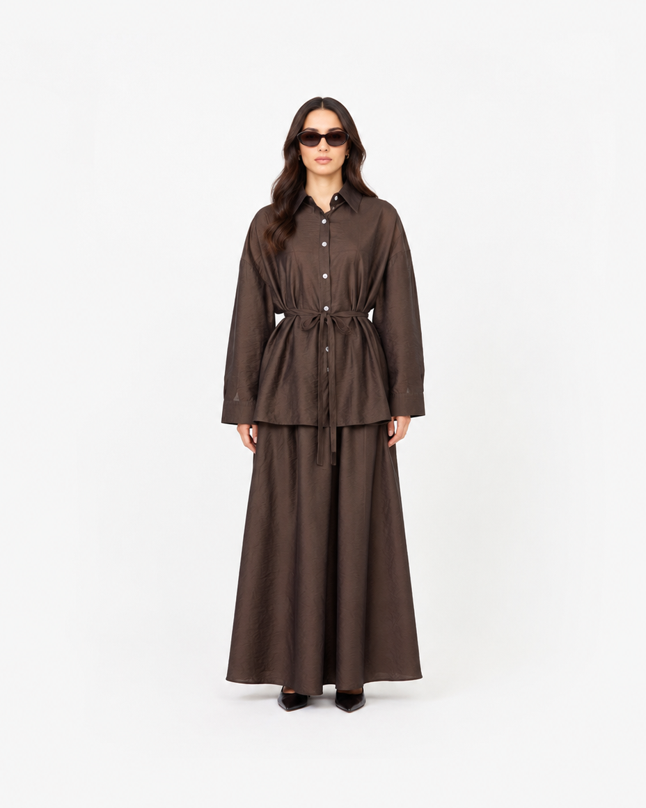 A model wearing a dark brown two-piece outfit consisting of a long-sleeve button-front shirt jacket with a tie waist and a matching long skirt, standing against a plain white studio background.