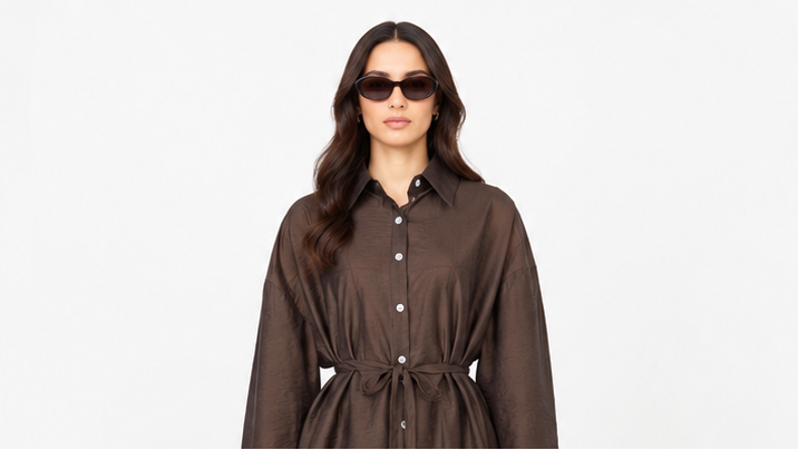 A woman modeling a dark brown long-sleeve button-up shirt dress with a tie waist, photographed against a plain white background.