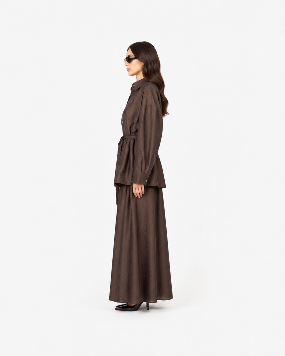A woman in side profile wearing a dark brown belted long-sleeve jacket and matching wide-leg skirt against a plain white studio background.