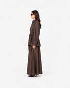 A woman in side profile wearing a dark brown belted long-sleeve jacket and matching wide-leg skirt against a plain white studio background.