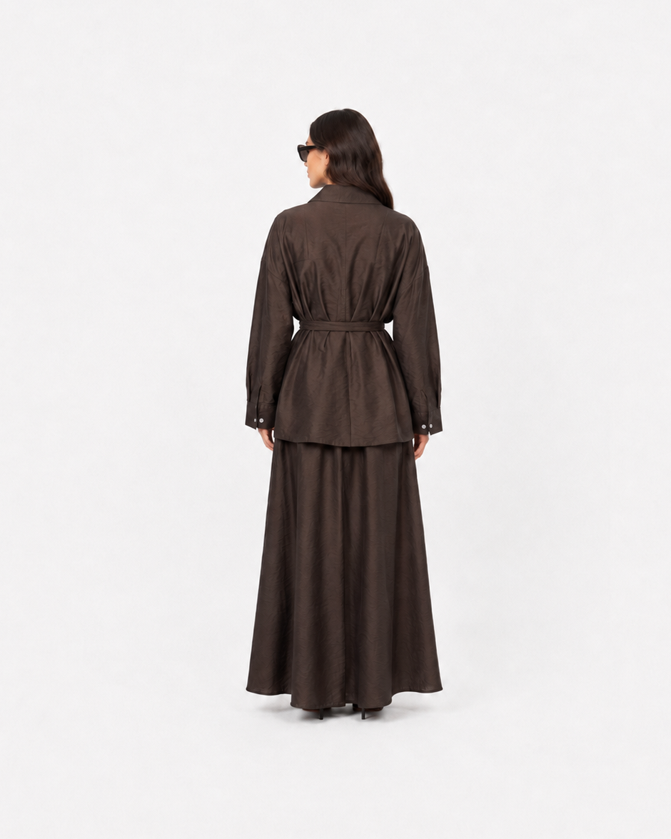 A model viewed from behind wearing a dark brown long-sleeved jacket with a matching waist tie and a long, flowing skirt against a plain white studio background.