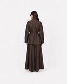 A model viewed from behind wearing a dark brown long-sleeved jacket with a matching waist tie and a long, flowing skirt against a plain white studio background.