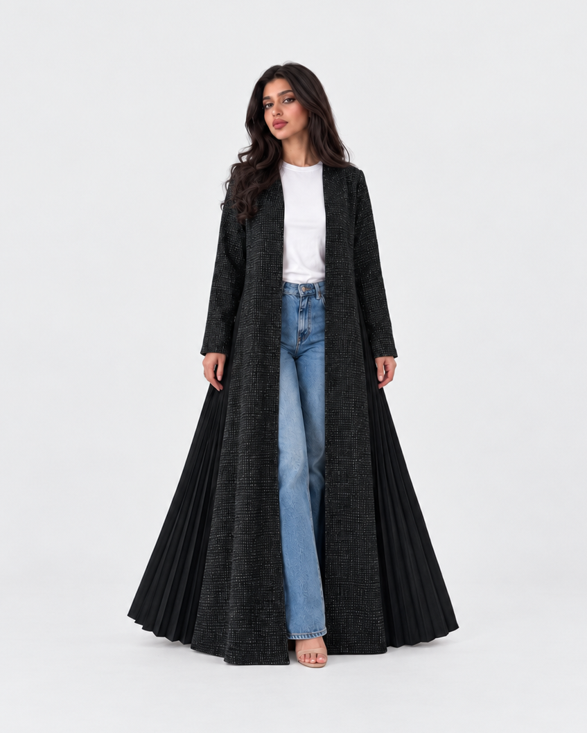 A woman modeling a long, black, open-front pleated coat over a white top and light blue jeans, standing against a seamless white studio background.