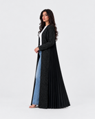 A woman modeling a long black pleated abaya with a textured outer layer, worn over a white top and light blue jeans, against a plain white studio background.