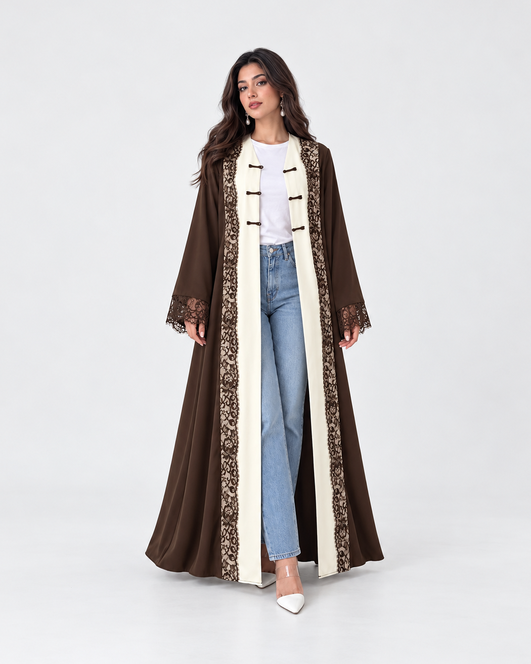 A woman modeling a long brown abaya with cream panels and intricate lace trim, styled with a white t-shirt, light blue jeans, and white pointed-toe heels against a plain white studio background.