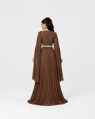 A woman modeling a floor-length brown pleated gown from the back, featuring long bell sleeves and a thin silver belt at the waist, set against a plain light gray studio background.