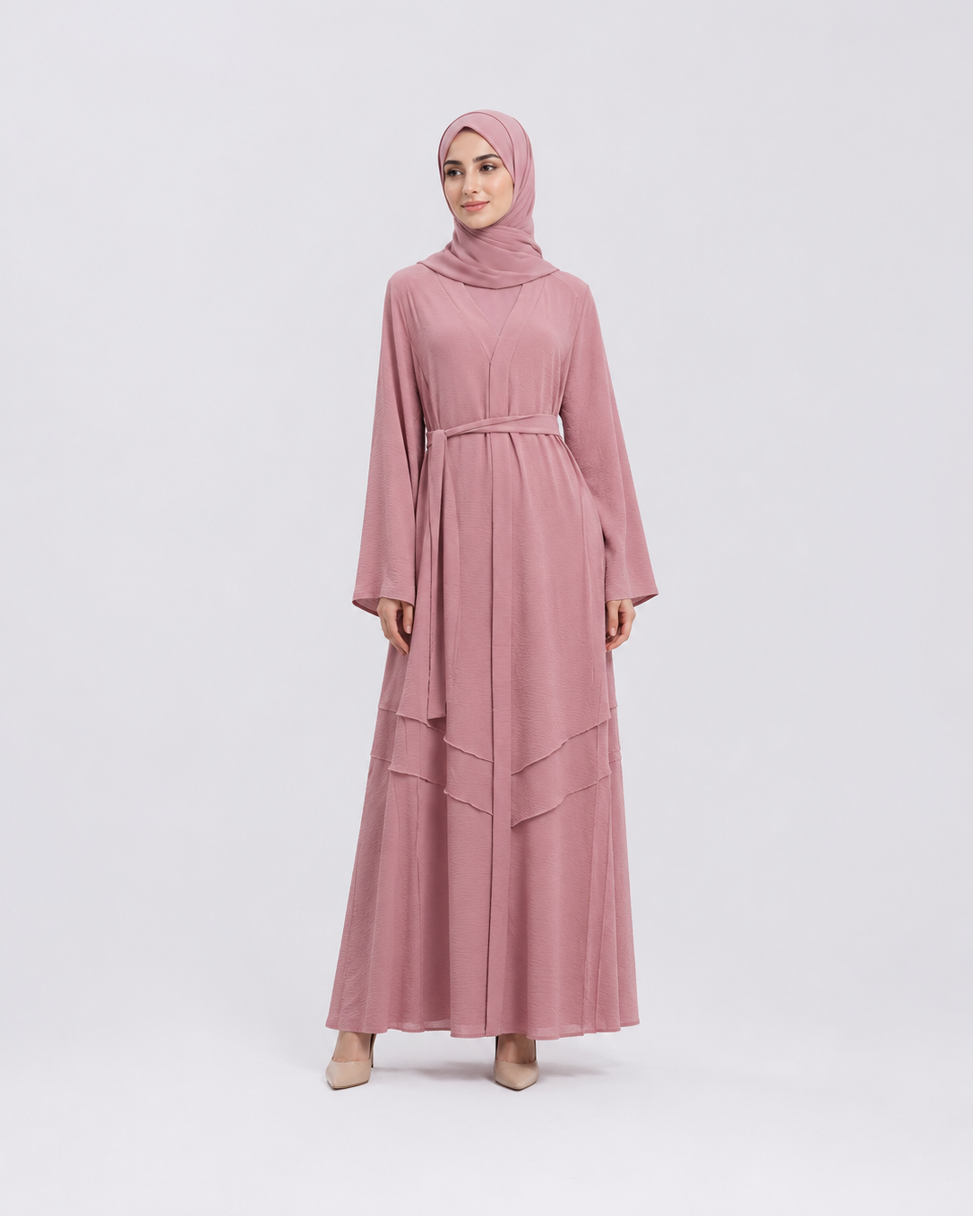 A woman modeling a dusty pink long-sleeved maxi abaya with a matching hijab, standing against a light gray studio background.