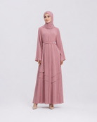 A woman modeling a dusty pink long-sleeved maxi abaya with a matching hijab, standing against a light gray studio background.