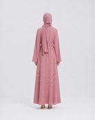 A woman viewed from the back wearing a long dusty pink abaya with a matching headscarf and a thin waist tie, standing against a light gray studio background.