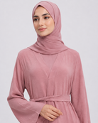 A woman wearing a dusty pink long-sleeve abaya with a matching waist tie belt and a coordinating hijab, photographed against a light gray studio background.