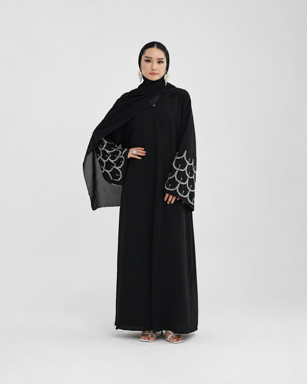 A woman modeling a long black abaya with wide sleeves featuring a white scalloped pattern, paired with a matching black hijab, against a light gray studio background.