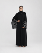 A woman modeling a long black abaya with wide sleeves featuring a white scalloped pattern, paired with a matching black hijab, against a light gray studio background.