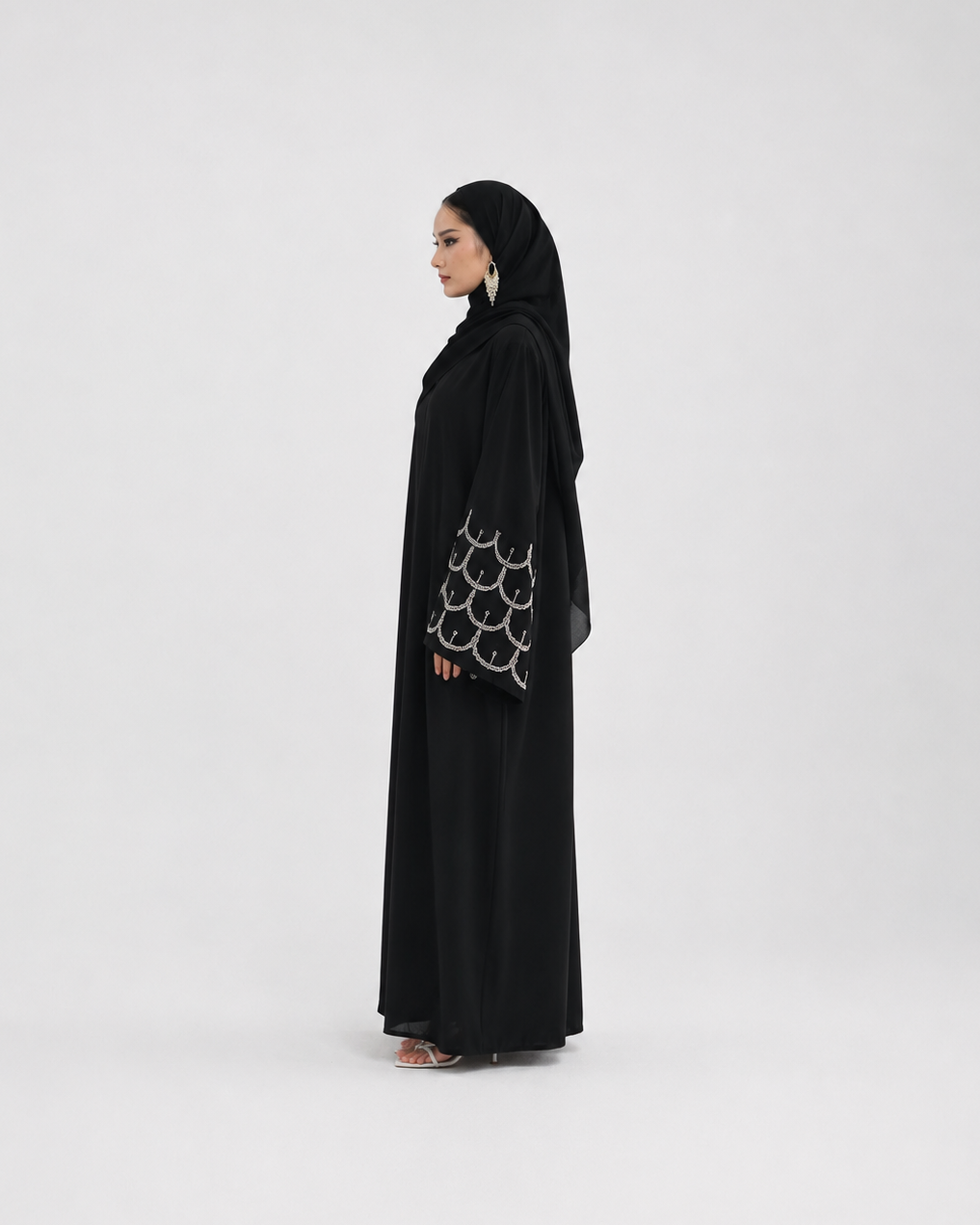 A woman wearing a black abaya with a matching black hijab, standing in profile against a plain light gray studio background. The abaya has a distinctive silver-tone circular pattern on the sleeve.