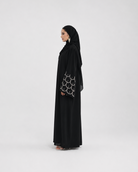 A woman wearing a black abaya with a matching black hijab, standing in profile against a plain light gray studio background. The abaya has a distinctive silver-tone circular pattern on the sleeve.