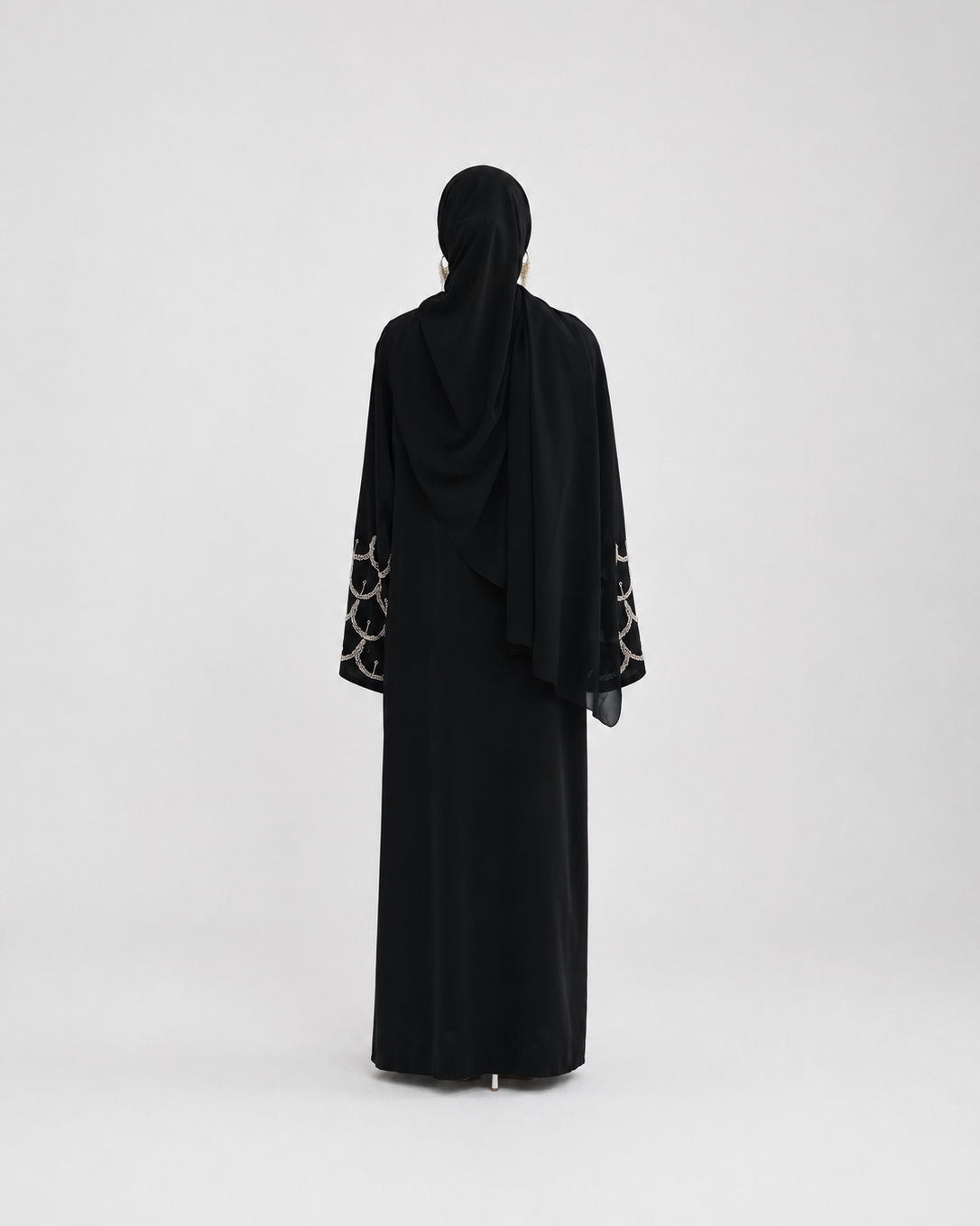 A woman viewed from behind wearing a black abaya with patterned sheer sleeves and a matching black hijab, standing against a plain light gray studio background.