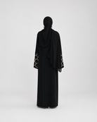 A woman viewed from behind wearing a black abaya with patterned sheer sleeves and a matching black hijab, standing against a plain light gray studio background.