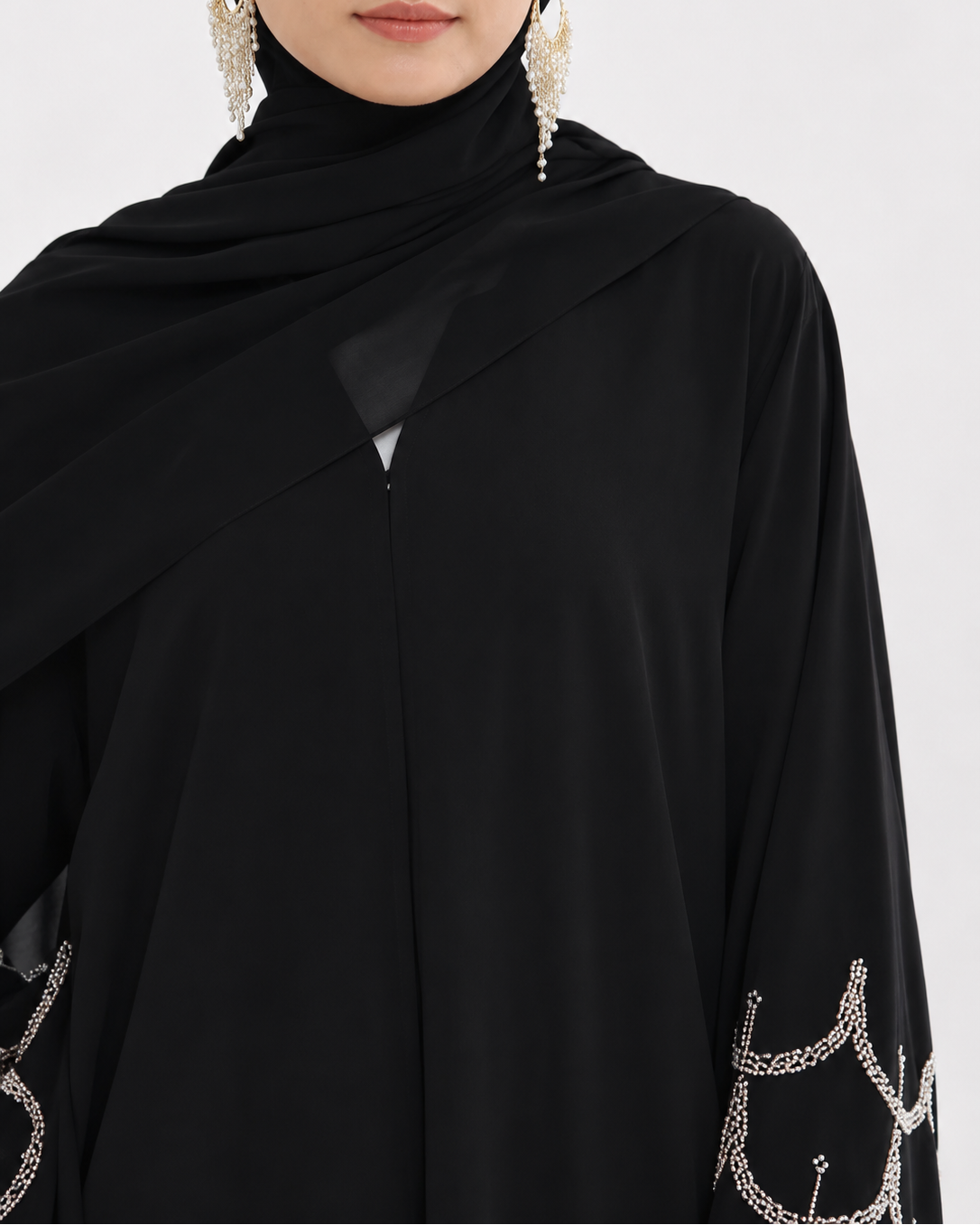 A close-up of a model wearing a black high-neck abaya with draped sleeves and silver chain embroidery on the cuffs, paired with a black hijab and dangling silver earrings.