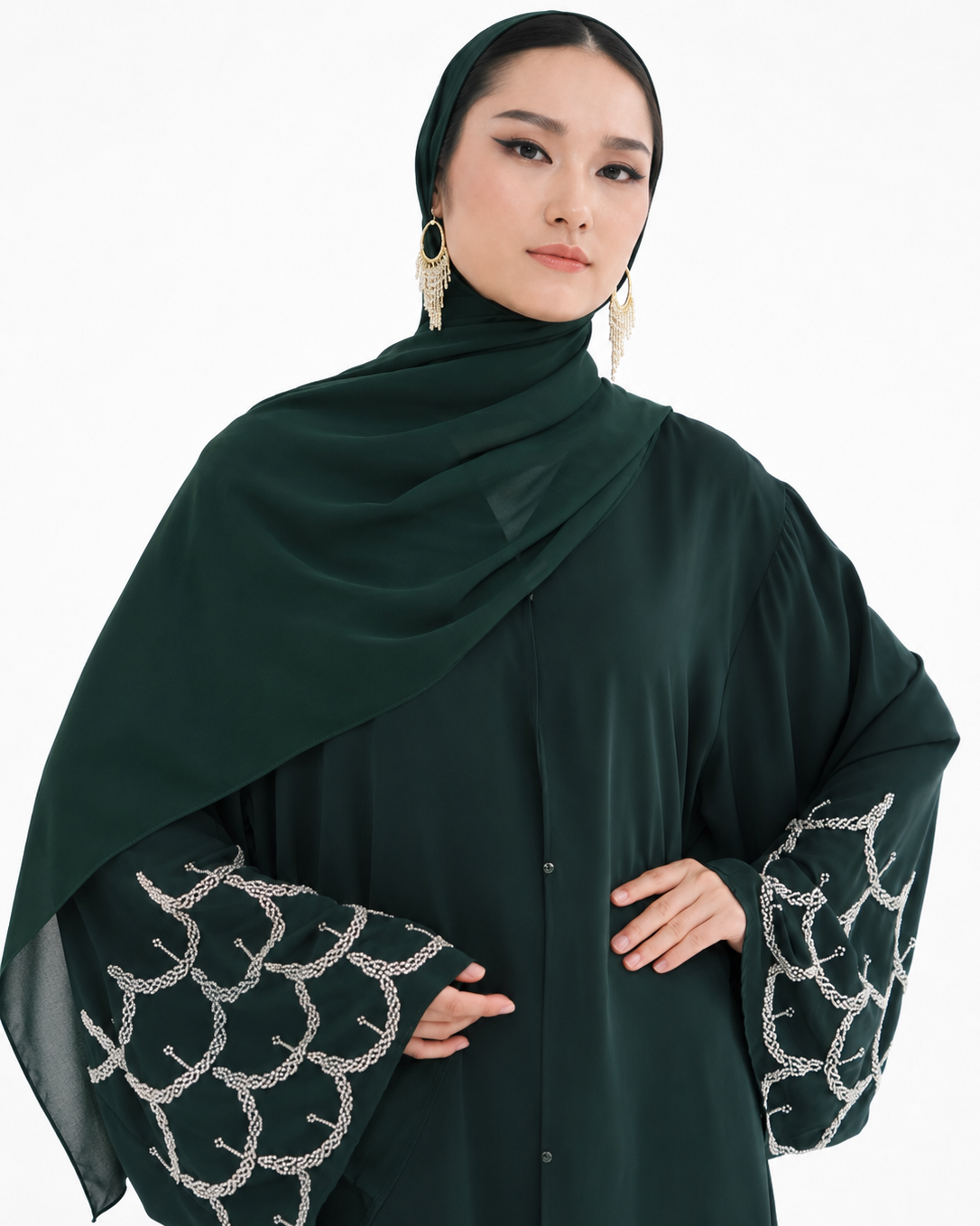 A woman wearing a dark forest green modest outfit featuring a draped hijab, a long-sleeved button-front abaya, and wide bell sleeves with silver chain-link embroidery, set against a plain white background.