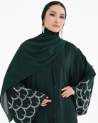 A woman wearing a dark forest green modest outfit featuring a draped hijab, a long-sleeved button-front abaya, and wide bell sleeves with silver chain-link embroidery, set against a plain white background.