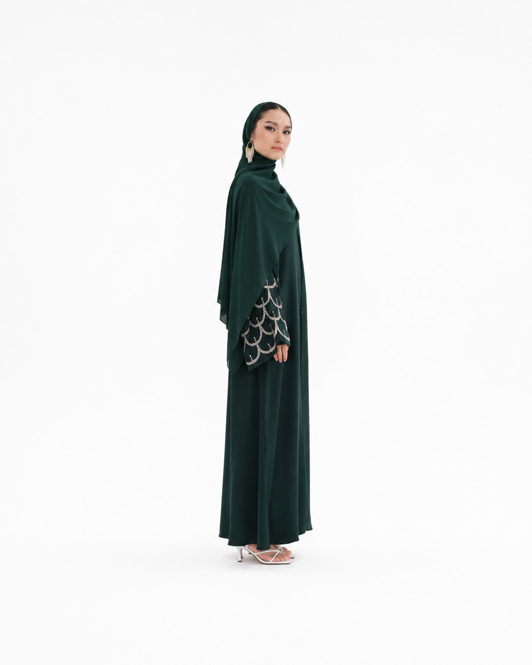 A woman modeling a dark green modest outfit consisting of a long abaya with patterned sleeves and a matching hijab, standing against a plain white studio background.