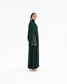 A woman modeling a dark green modest outfit consisting of a long abaya with patterned sleeves and a matching hijab, standing against a plain white studio background.