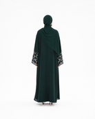 A woman viewed from behind wearing a dark forest green abaya with a matching hijab. The abaya features a draped design with patterned black and white circular lace cuffs on the sleeves.