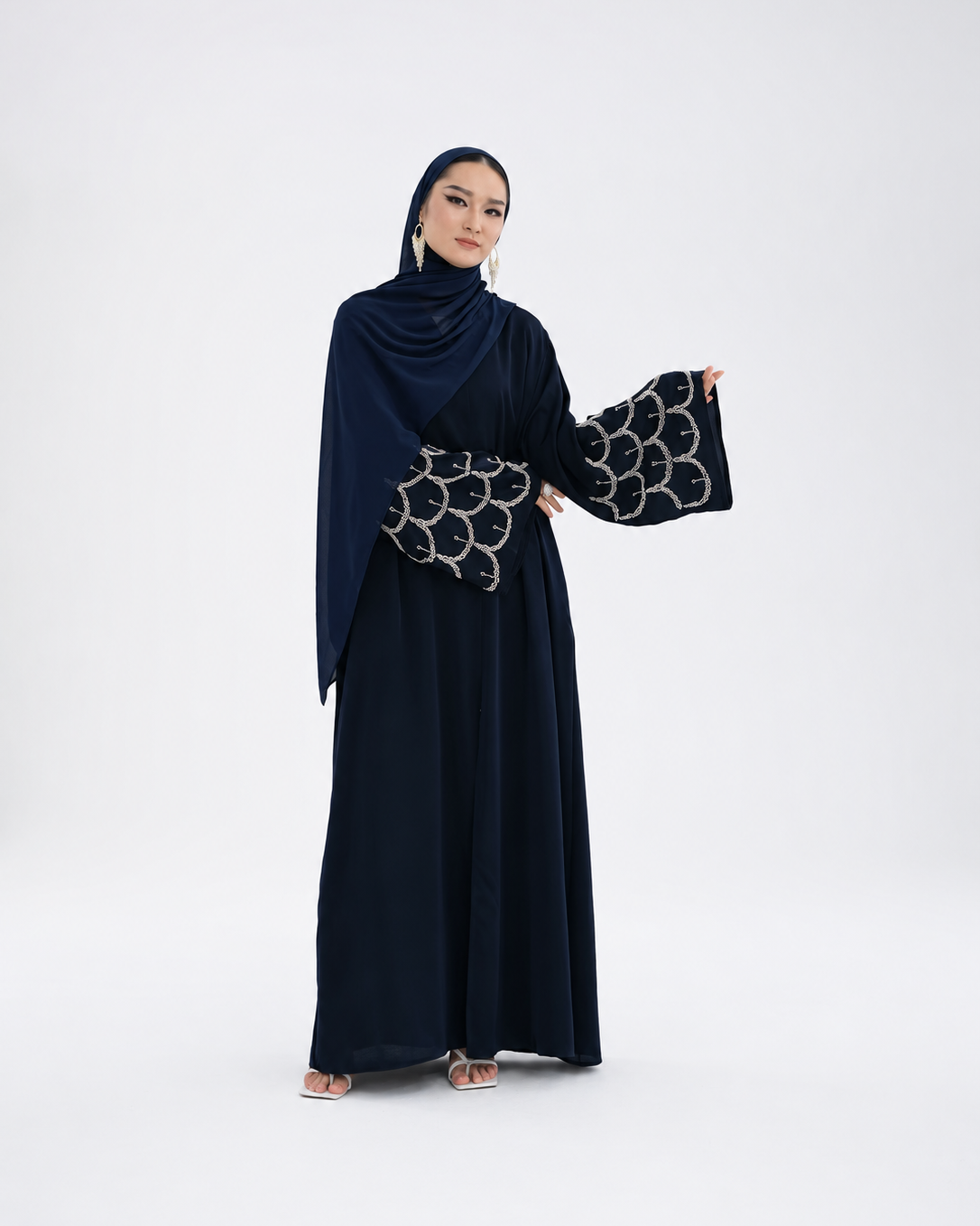 A woman modeling a dark navy blue abaya dress with wide sleeves featuring a silver geometric pattern, paired with a matching draped hijab, against a plain light gray studio background.