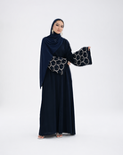 A woman modeling a dark navy blue abaya dress with wide sleeves featuring a silver geometric pattern, paired with a matching draped hijab, against a plain light gray studio background.