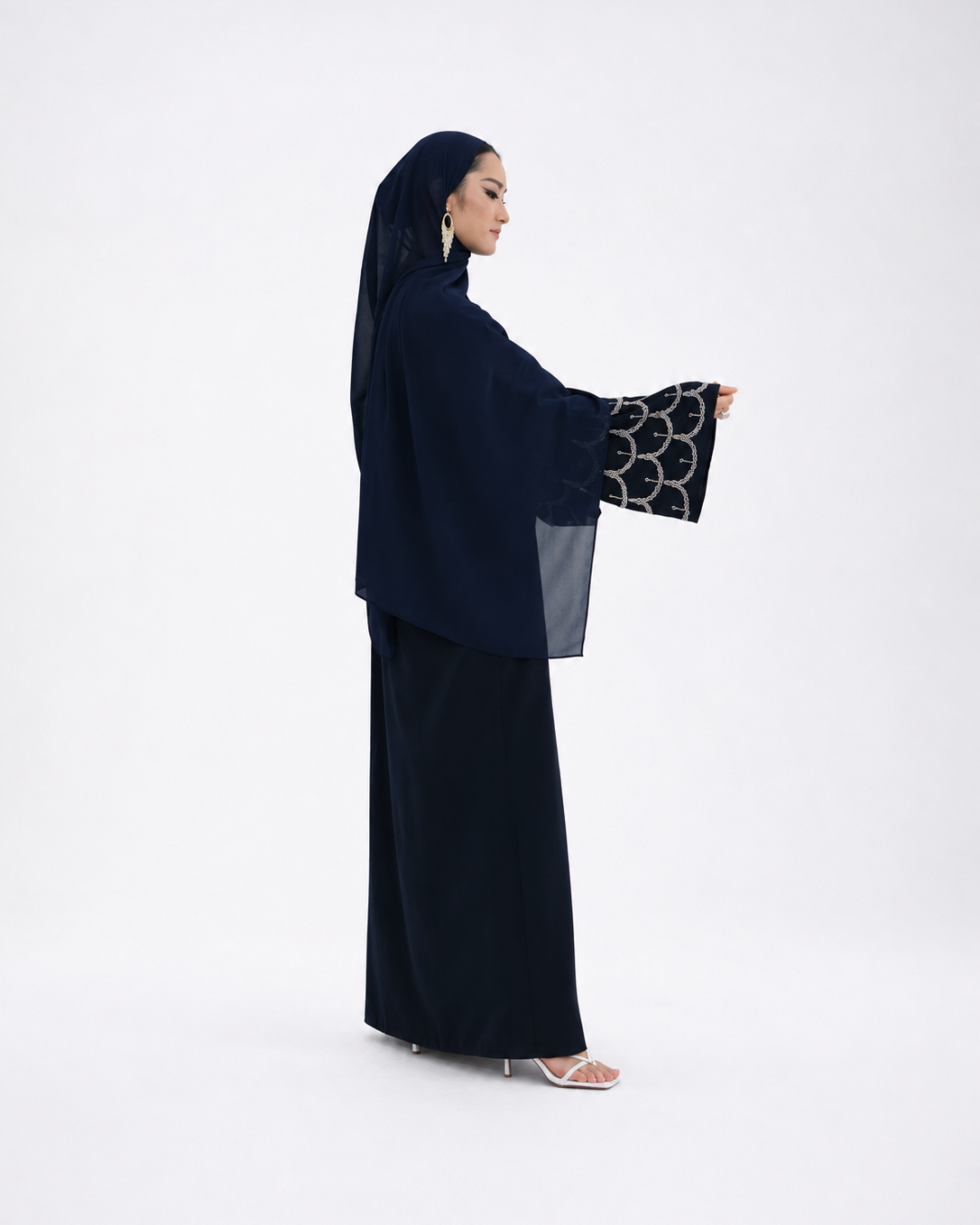A woman modeling a dark navy abaya with wide sleeves featuring a black and white circular pattern, paired with a matching navy hijab and skirt, set against a plain white studio background.
