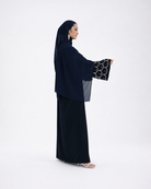 A woman modeling a dark navy abaya with wide sleeves featuring a black and white circular pattern, paired with a matching navy hijab and skirt, set against a plain white studio background.