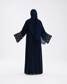 A woman wearing a dark navy abaya and matching hijab, shown from the back against a plain white studio background. The abaya has wide sleeves with a black and white circular patterned trim at the cuffs.