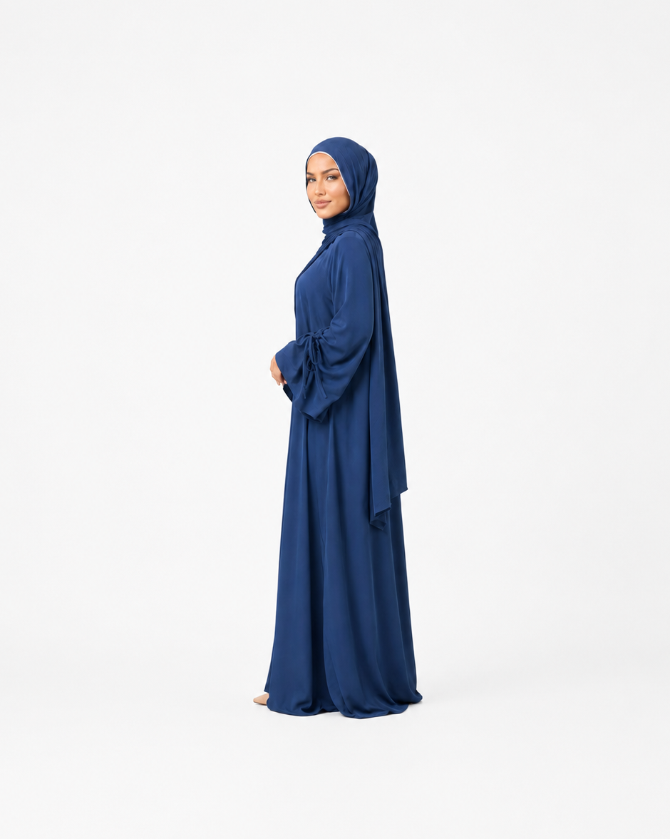 A woman wearing a long navy blue abaya and matching hijab stands in profile against a plain white studio background.