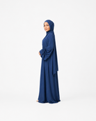 A woman wearing a long navy blue abaya and matching hijab stands in profile against a plain white studio background.