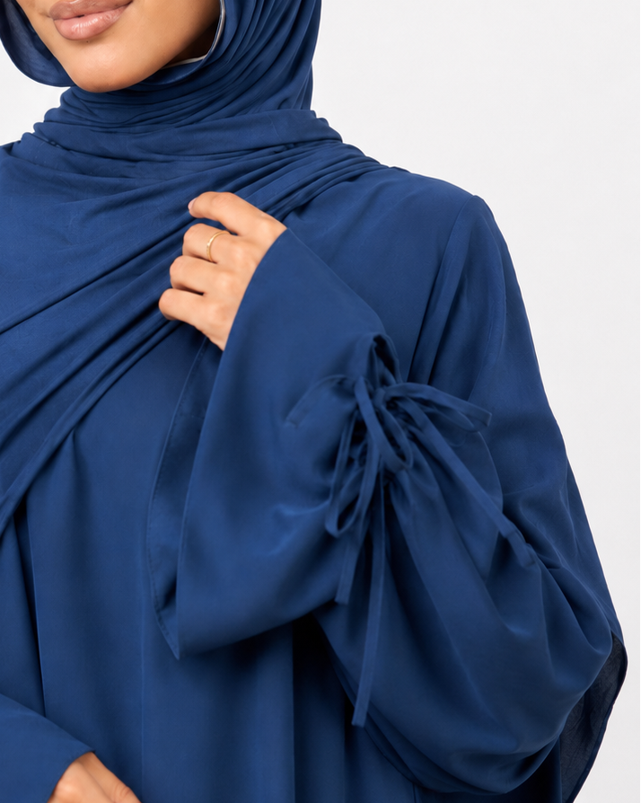 A woman wearing a navy blue modest outfit consisting of a draped hijab and a matching long-sleeve abaya with tie details at the cuffs, photographed against a light gray studio background.