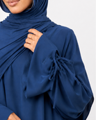 A woman wearing a navy blue modest outfit consisting of a draped hijab and a matching long-sleeve abaya with tie details at the cuffs, photographed against a light gray studio background.