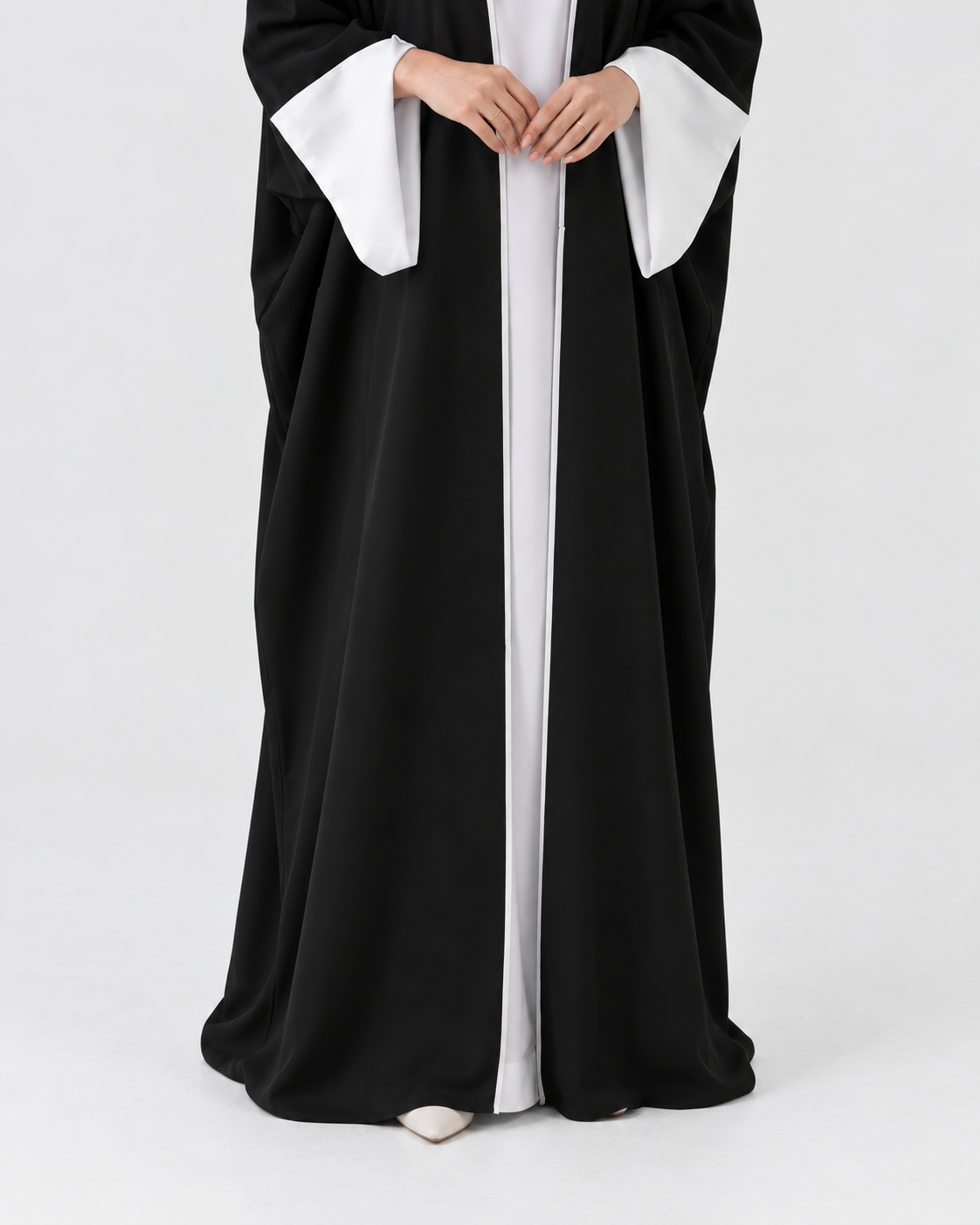 A person wearing a long black robe with wide white sleeves and a white front panel, standing against a plain light gray studio background.