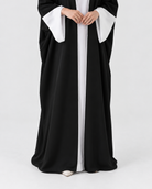 A person wearing a long black robe with wide white sleeves and a white front panel, standing against a plain light gray studio background.