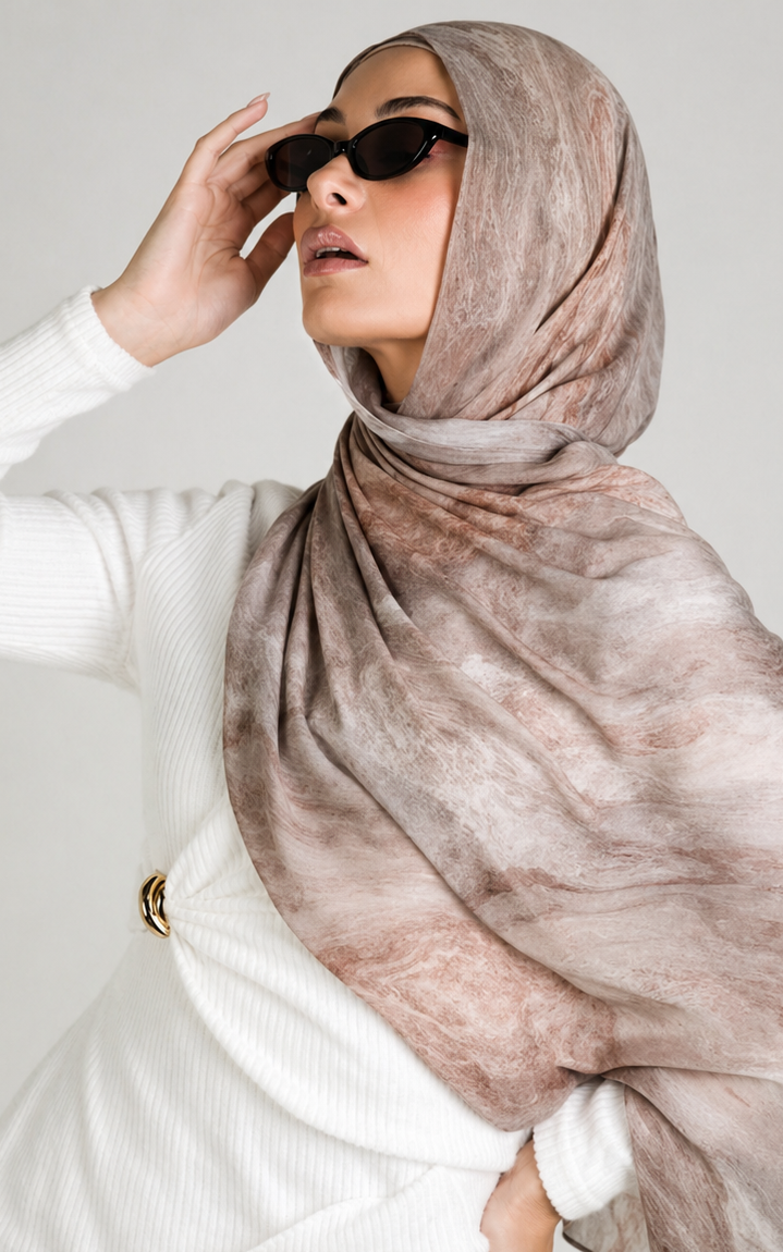 A woman wearing a large, lightweight hijab with a soft tie-dye pattern in dusty pink, taupe, and beige. She is wearing a white ribbed sweater and black sunglasses, posing against a light gray studio background.