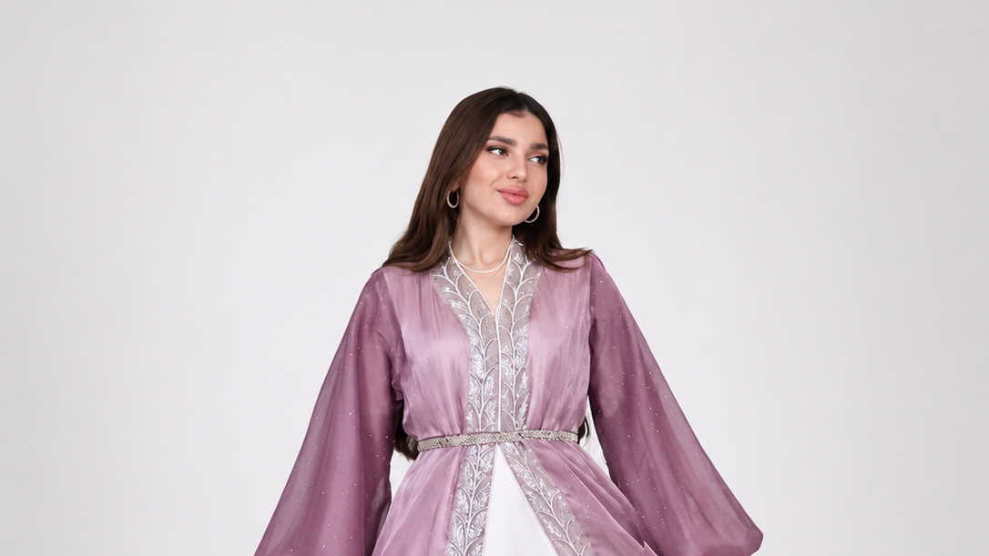 A woman wearing a lavender-purple traditional long-sleeve dress with silver embroidery and a white underlayer, standing against a plain light gray background.
